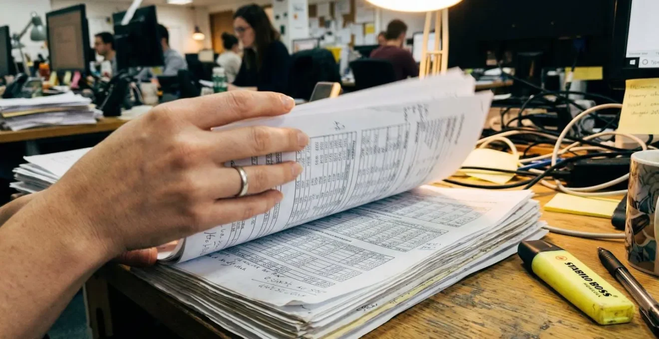 Mains feuilletant des pages imprimées avec tableaux de chiffres, surligneur jaune posé sur le côté